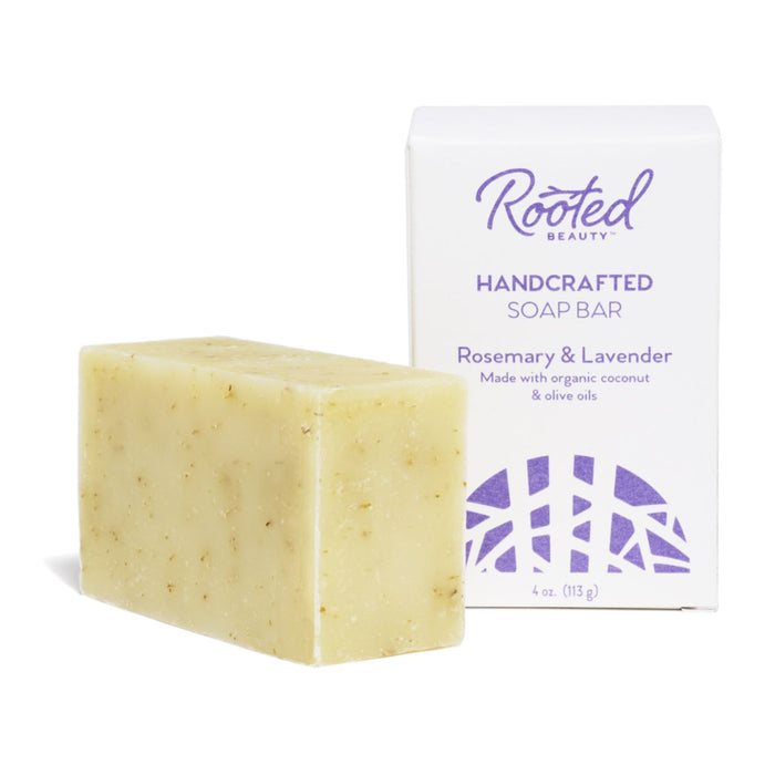 A beige soap bar, textured and unwrapped, rests beside its box labeled Rooted Beauty Bar Soap. The packaging highlights a cruelty-free, moisturizing formula featuring organic coconut oil and oatmeal, with Rosemary & Lavender printed in purple text.