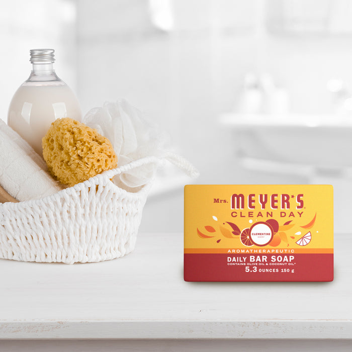 Mrs. Meyers Bar Soap, Leaping Bunny certified with garden-inspired packaging featuring leaves and an orange design, is displayed on a bathroom counter next to a white basket containing a sponge, towels, and a bottle that looks like glass.