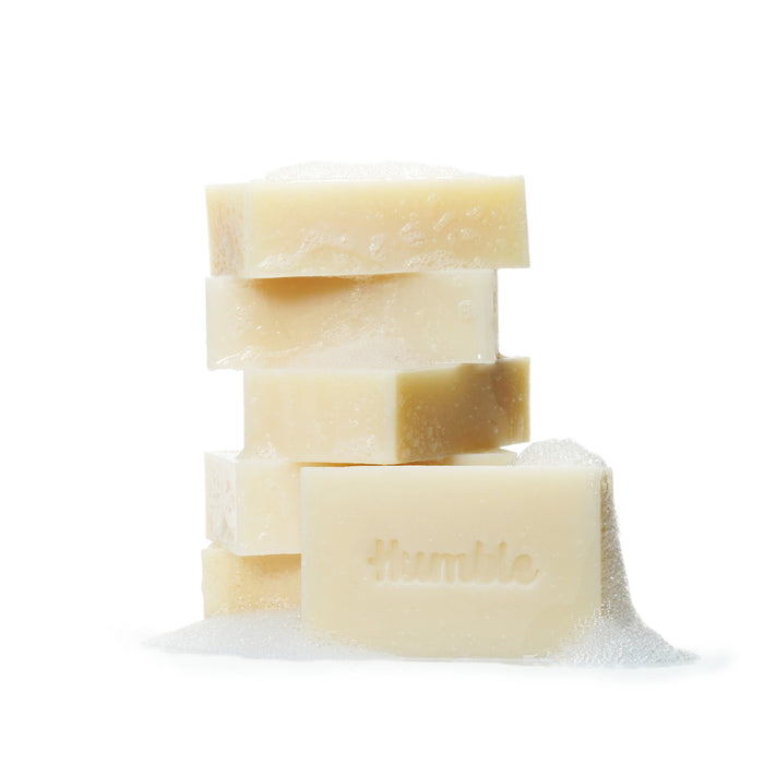 A stack of beige Humble Brands Bar Soaps sits on a white background, with bubbles decorating the top and sides. The front soap bears humble, highlighting its eco-friendly nature and natural oils.