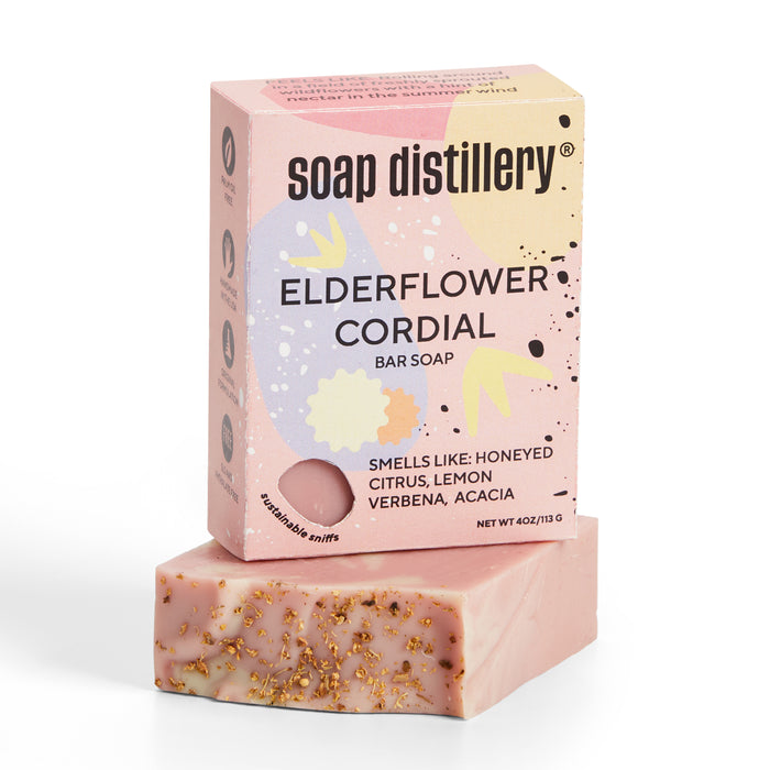 A pink bar of Soap Distillerys Elderflower Cordial Bar Soap, featuring dried flowers, rests before its matching box. The package highlights scents of honeyed citrus, lemon verbena, acacia and showcases decorative graphics. It is palm oil free.