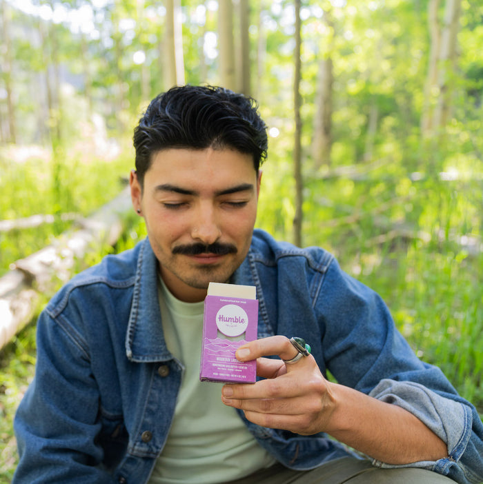A person with dark hair and a mustache, in a denim jacket and light green shirt, sits outdoors in a lush, green forest. They gaze at an eco-friendly Humble Brands Bar Soap, handcrafted from natural oils, held in their hand.