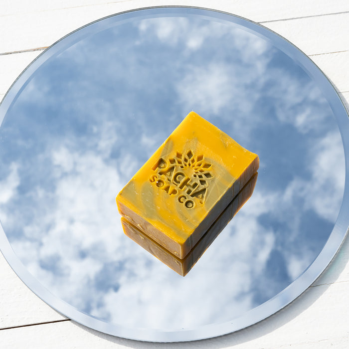A Pacha Soap Co. bar of brown and orange soap, infused with Clarifying Charcoal, is elegantly reflected against a cloudy blue sky on a round mirror over a white wooden surface.