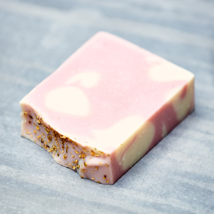 The Soap Distillery Bar Soap, rectangular with pink and white swirls, aromatic scents, and gold flecks on one edge, elegantly rests on a gray surface.