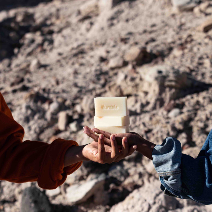 Against a rocky background, two hands exchange Humble Brands Bar Soap. One person wears an orange sleeve, the other a denim jacket. This handcrafted soap, enriched with natural oils, is an eco-friendly choice for conscientious consumers.