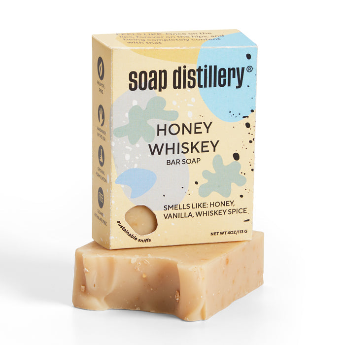 A bar of Bar Soap by Soap Distillery rests on a white background, exuding honey, vanilla, and whiskey spice aromas. The fragrance comes in beige, blue, and olive green packaging. This palm oil-free soap features a light brown color.