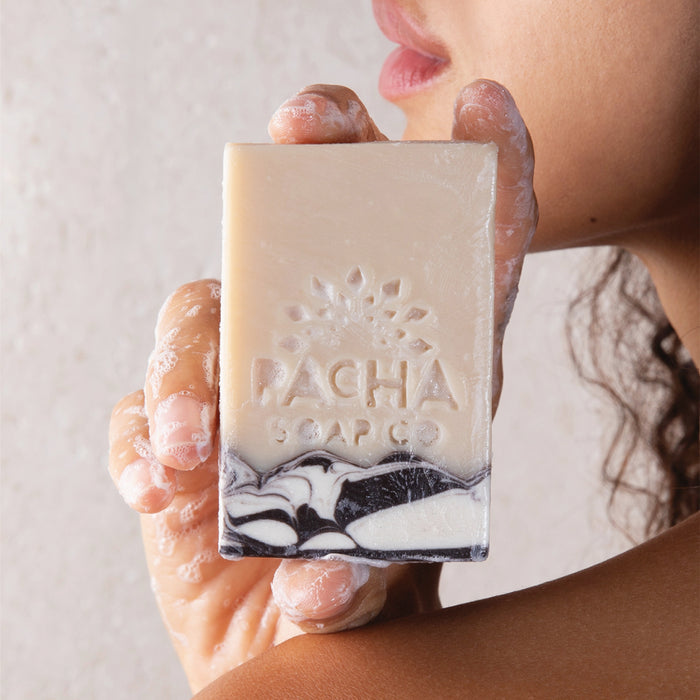 A person holds Pacha Soap Co. Bar Soap near their face, with nourishing oils and soap suds on their hand and shoulder, set against a neutral background.
