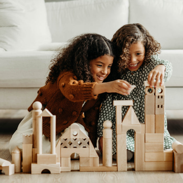 Two curly-haired children happily play on a wooden floor with the HABA Basic Building Blocks - Extra Large Wooden Starter Set, building towers and arches in a cozy, bright living room with a white sofa.
