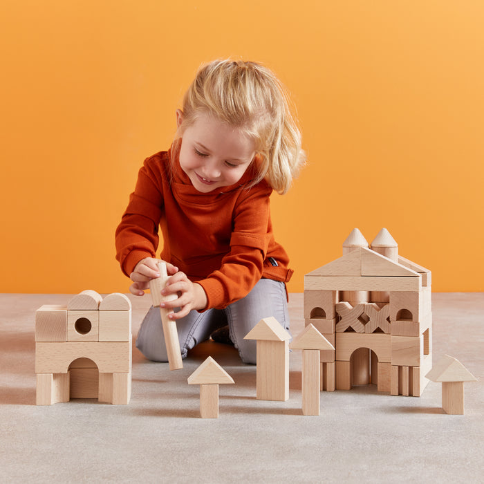 A blonde-haired child wearing an orange sweater and jeans creatively builds structures on the floor with HABAs Basic Building Blocks - Extra Large Wooden Starter Set, set against a vibrant orange background.