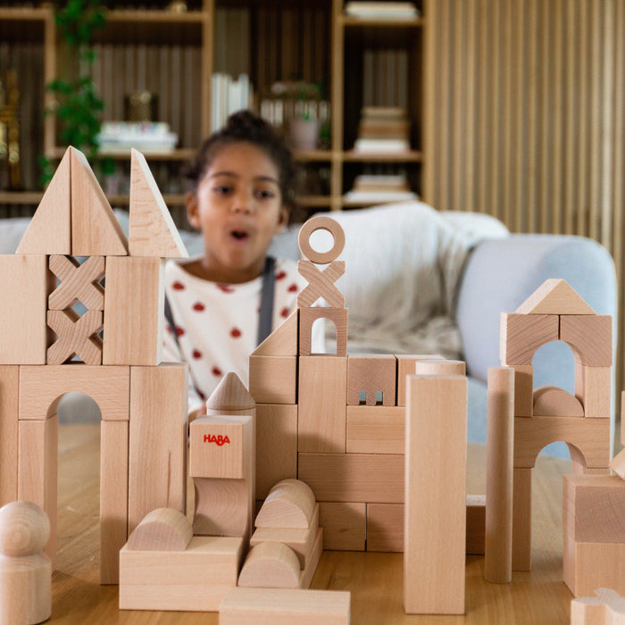A child with a surprised expression sits behind a table filled with HABAs Basic Building Blocks - Extra Large Wooden Starter Set, made from sustainably sourced wood, showcasing creative play through various shapes and structures in a cozy room with shelves and a couch.