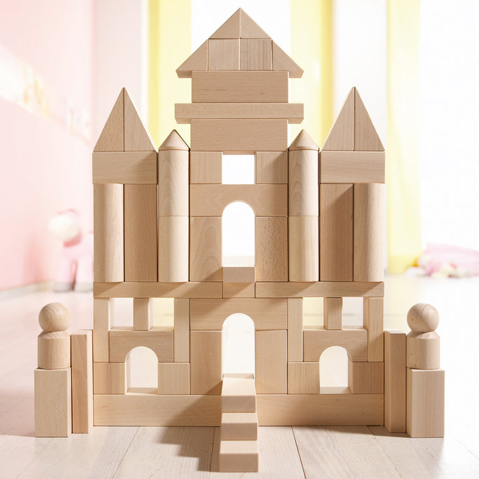A symmetrical castle made from HABAs Basic Building Blocks - Large Starter Set features towers, arches, and a central staircase. Two block figures stand at the entrance, set against a softly lit background with pastel hues, highlighting the charm of sustainable toys that inspire child development.