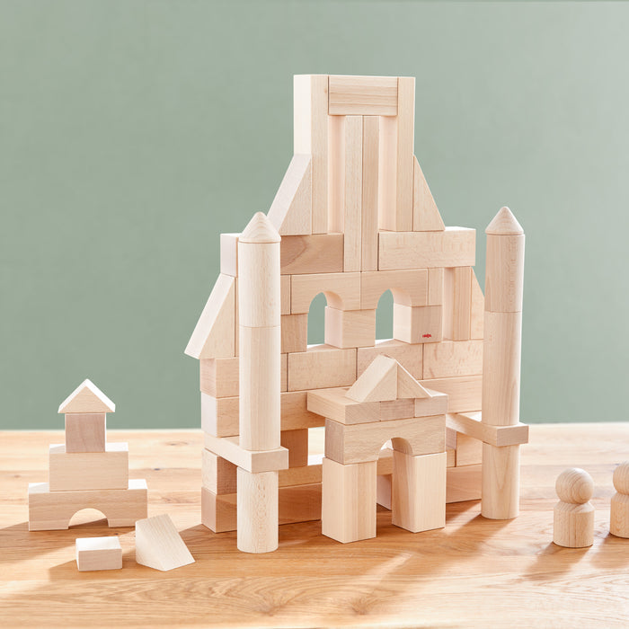 HABAs Basic Building Blocks - Large Starter Set is stacked into a house-like structure with towers and arches. This setup sits on a wooden table against a light green background, accompanied by two wooden peg figures.