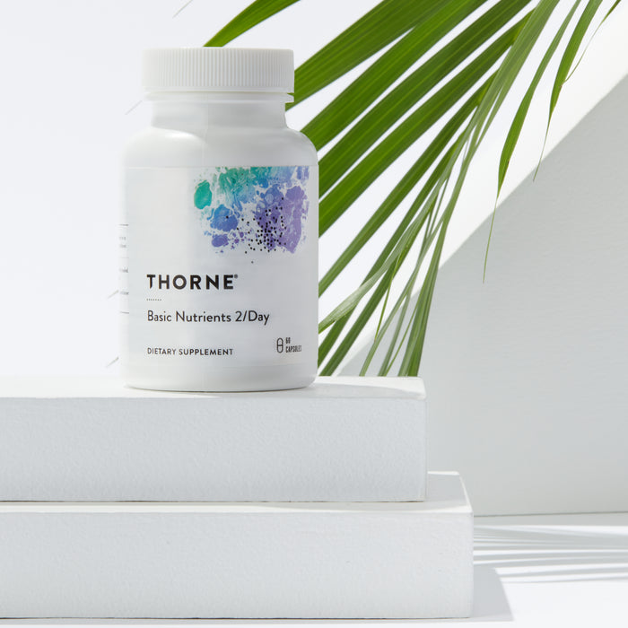 A Thorne Basic Nutrients 2/Day bottle rests on white blocks, its label showcasing colorful abstract art. Sunlit green palm leaves cast shadows in the background, highlighting the supplements role in offering nutritional support.