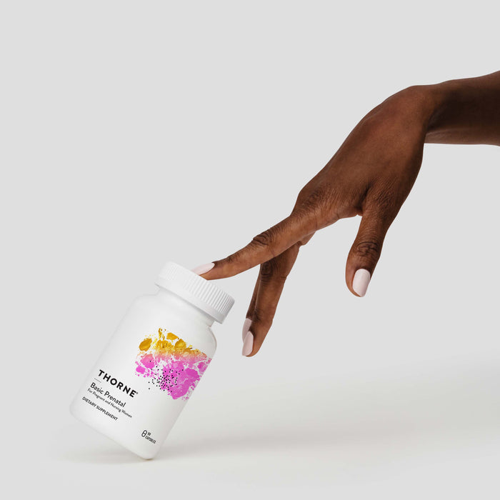 A hand with light-colored nails gently tilts a bottle of Thornes Basic Prenatal, a daily womens multivitamin, against a plain background. The bottle showcases a colorful pink, yellow, and purple splash design to support baby development effectively.