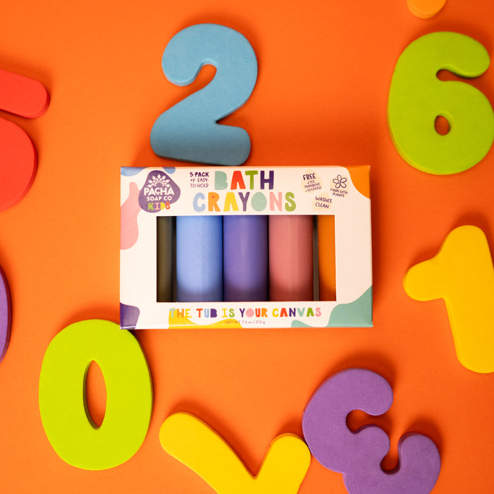 Pacha Soap Co.s Bath Crayon box features non-toxic, natural colorants in white packaging with vibrant light blue, purple, and pink colors. Its set against an orange background with colorful foam numbers: 2, 6, 0, and 5.