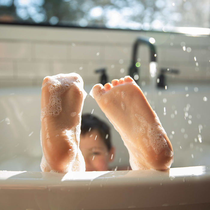 Two feet covered in suds playfully stick out of a non-toxic bathtub while Pacha Soap Co.s colorful Bath Crayons add a playful touch. A childs head is barely visible behind the tub, water droplets glistening as sunlight filters through the window, creating a cheerful scene.