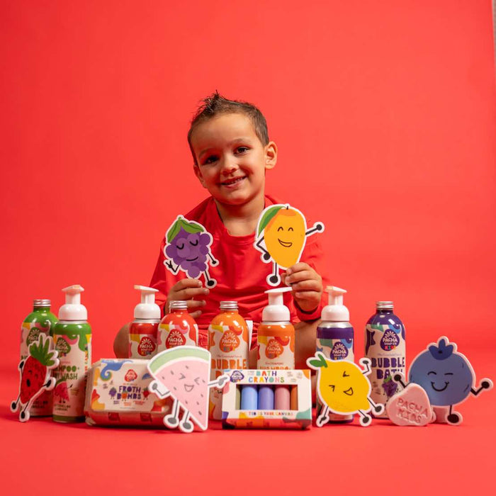 A smiling child sits against a red background, holding cartoon fruit characters. In front, Pacha Soap Co.s non-toxic Bath Crayons and colorful bath products with playful fruit faces are displayed, made with natural colorants for safe and vibrant fun.