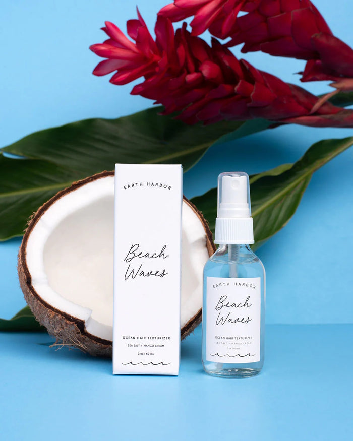 A spray bottle of BEACH WAVES Ocean Hair Texturizer by Earth Harbor stands next to its box, with a halved coconut and large green leaves. Red tropical flowers set against a light blue backdrop capture the vibrant essence of mango sea salt spray in a breezy beach style scene.
