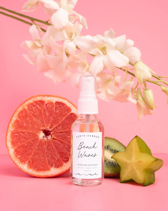 A spray bottle labeled BEACH WAVES Ocean Hair Texturizer from Earth Harbor exudes beach style, nestled among a halved grapefruit, sliced kiwi, and starfruit on a pink background. White flowers elegantly frame the vegan concoction that captures a tropical escapes essence.
