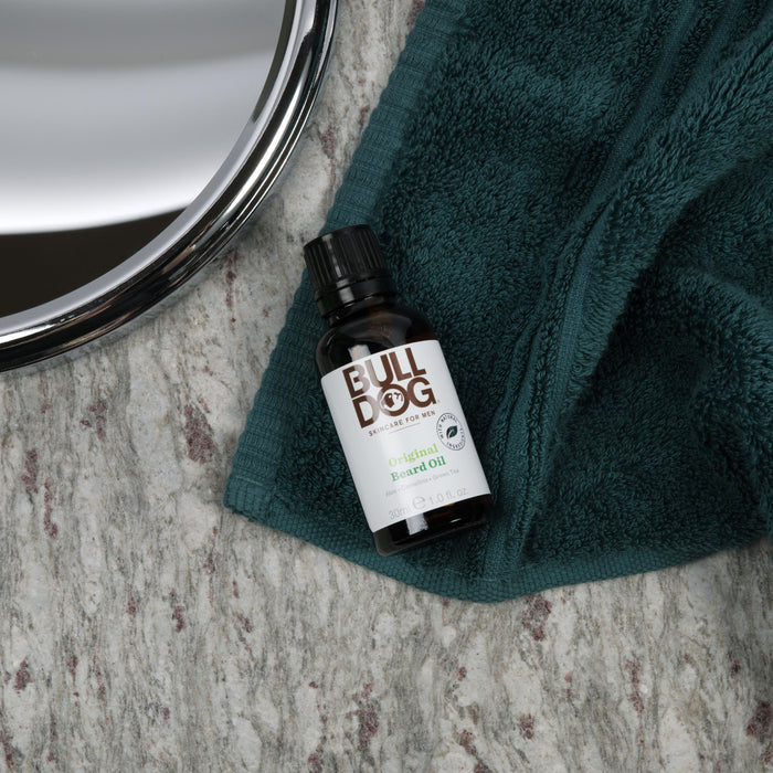 A bottle of Bulldog Beard Oil rests on a green towel next to a round mirror on a marbled surface.