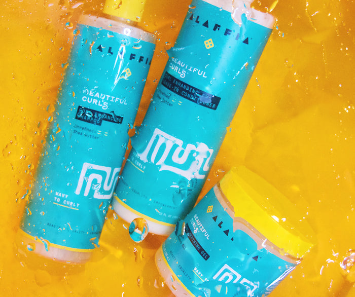 Three Alaffia Beautiful Curls products—Blue and yellow shampoo, leave-in conditioner, and curl defining gel—are set on a wet orange surface with water droplets, ideal for embracing natural curls and reducing frizz.