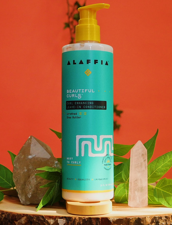 A turquoise bottle of Alaffias Beautiful Curls Curl Enhancing Leave-In Conditioner for wavy to curly hair, enriched with shea butter and a yellow pump, is placed on a wooden surface surrounded by green leaves, two crystals, and set against a vibrant red background.