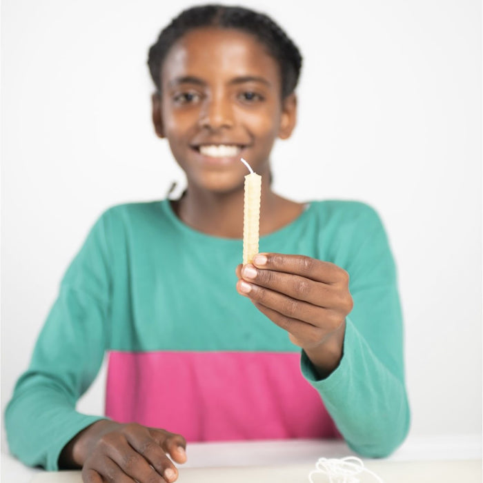 A person in a green and pink shirt smiles while holding a beeswax candle with a honeycomb pattern from the eco-kids Beeswax Honeycomb Candle Kit against a plain white background, with string on the table suggesting natural candle-making.