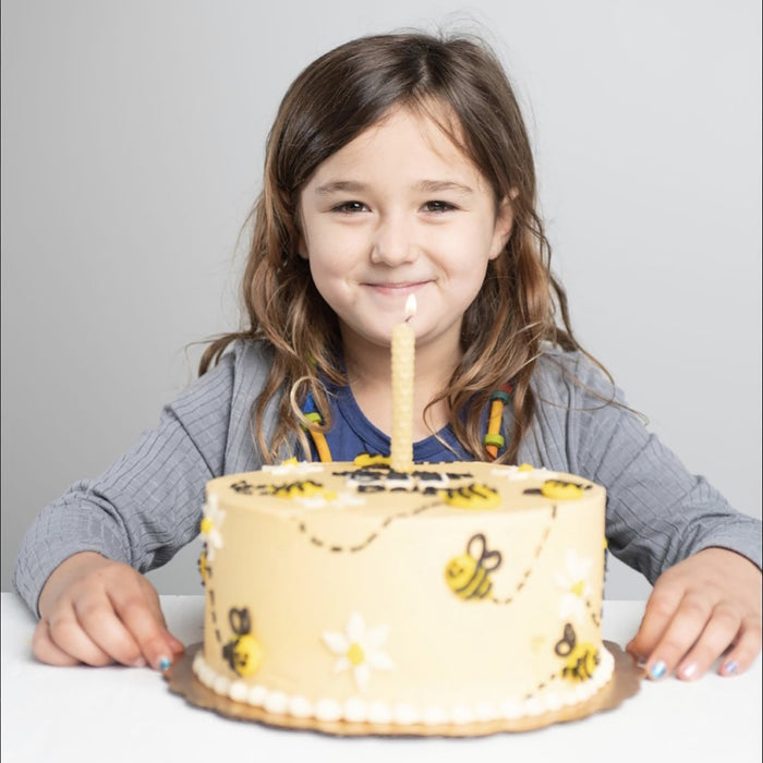 A young girl smiles behind a yellow cake with bees, flowers, and a single candle from the Beeswax Honeycomb Candle Kit by eco-kids. Shes wearing a gray top with long brown hair in front of a plain light gray background.
