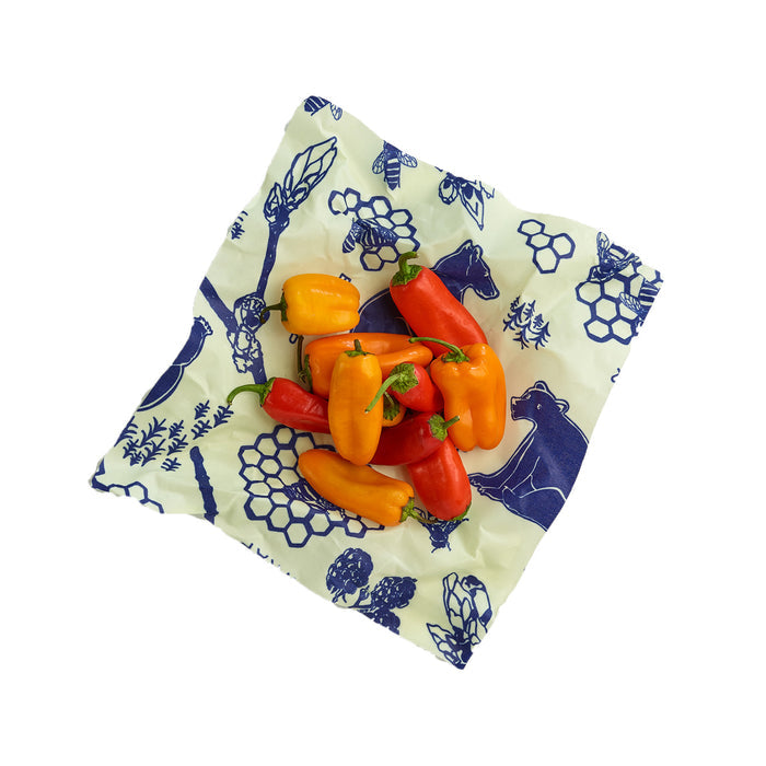 A cluster of small red and yellow peppers rests on a Bees Wrap Single Medium Wrap, featuring blue prints of bears, honeycombs, bees, and leaves on a white background. This charming beeswax wrap offers an eco-friendly touch to your kitchen storage.