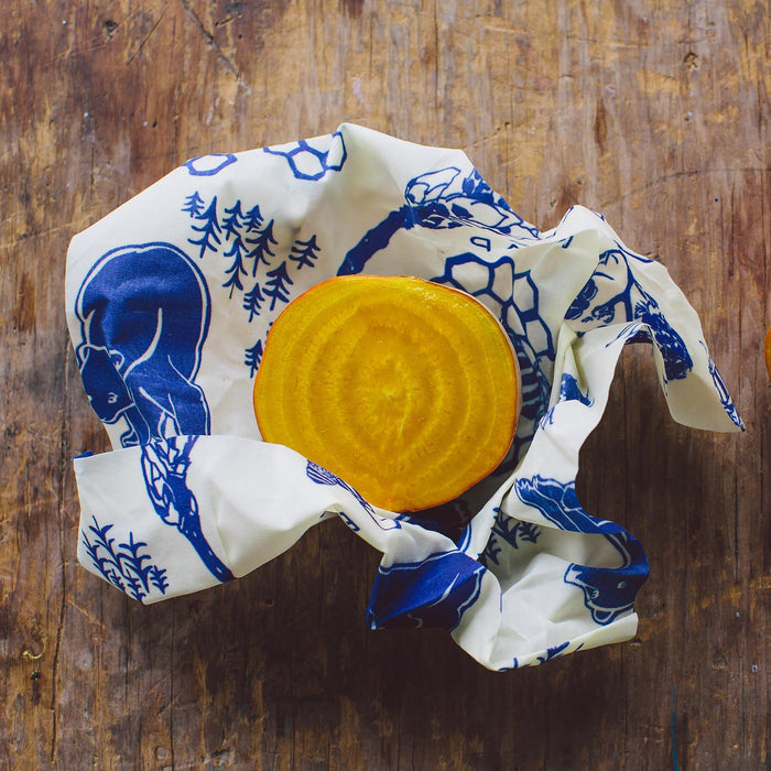 A halved golden beetroot lies on a wooden surface, wrapped in Bees Wraps Single Medium Wrap. The blue and white beeswax wrap features marine motifs like fish and seashells, providing a stylish and eco-friendly reusable food storage option.