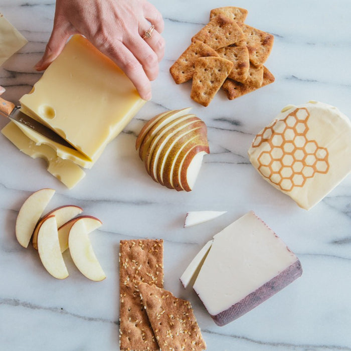 A hand slices cheese on marble, surrounded by apple and pear slices, crackers, and other cheeses—one wrapped in Bees Wrap Single Medium Wrap, a reusable alternative to plastic wrap with a honeycomb pattern.