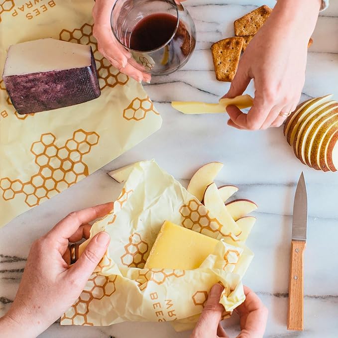 Hands prepare a snack with cheese, crackers, sliced pear, and a glass of red wine on marble. Some cheese is wrapped in Bee's Wrap Beeswax Wraps for Food - Large—a stylish, eco-friendly alternative to plastic wrap. A knife sits nearby.
