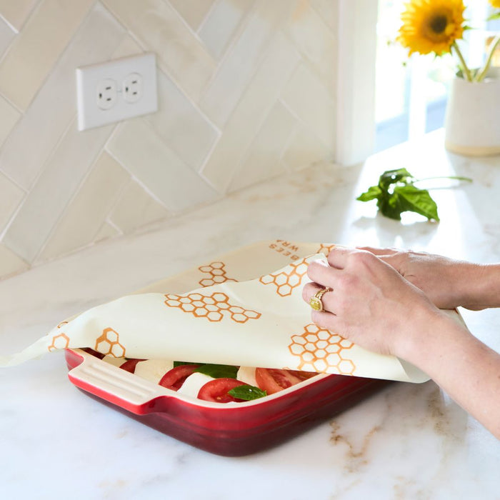 A person covers a red dish of sliced vegetables with Bee's Wrap Beeswax Wraps for Food - Large, a sustainable alternative to plastic wrap, on a kitchen counter. A sunflower in a vase and greens are visible in the background.