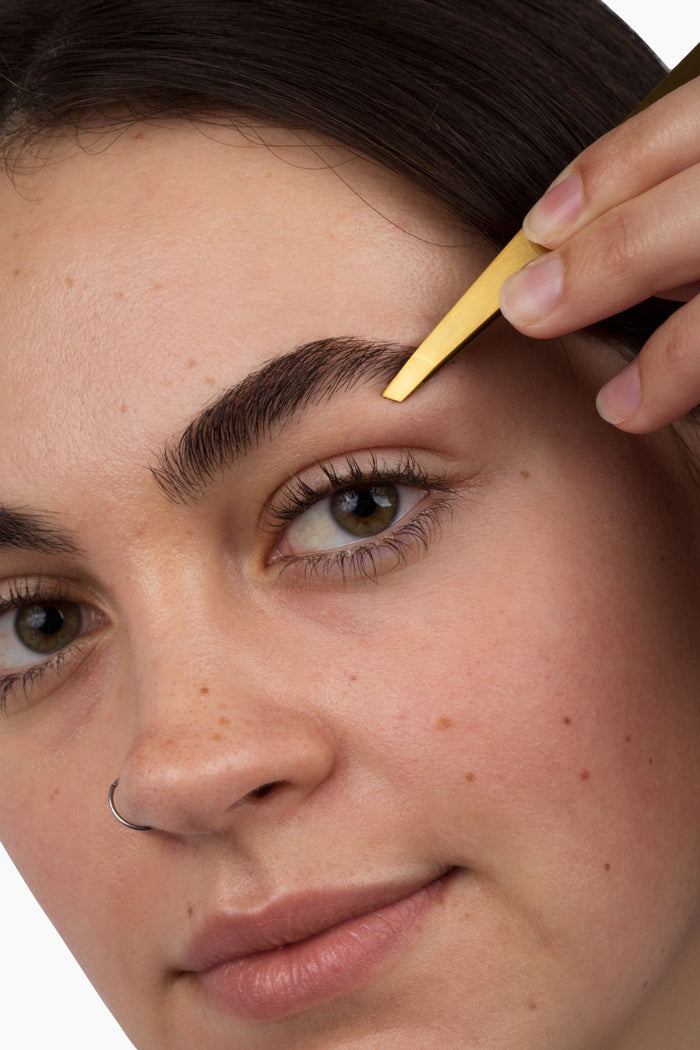 A close-up captures a person using the Terra Beauty Bars Beheld Eye Brow Kit, expertly shaping their eyebrows. The image focuses on the eye area, highlighting flawless skin and brow precision against a plain white background.