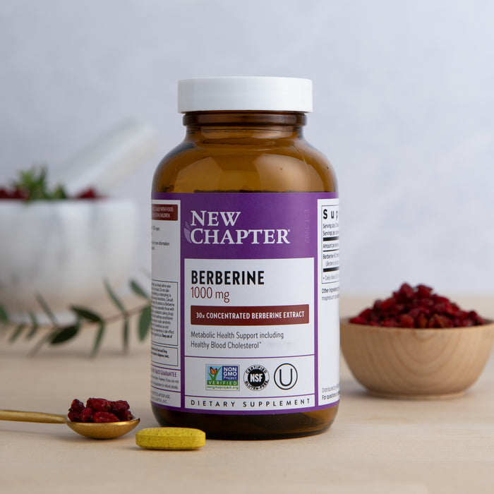 A 1000 mg Berberine bottle from New Chapter sits on a table, promoting metabolic and heart health. A yellow tablet lies next to a spoon with dried red berries, while more berries fill a wooden bowl in the background.