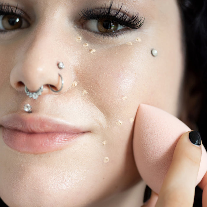 Close-up of a womans face applying makeup with the Ere Perez Bio All-Beauty Sponge, showcasing blending foundation on her cheek. She sports multiple nose and lip piercings and long eyelashes against a blurred background.