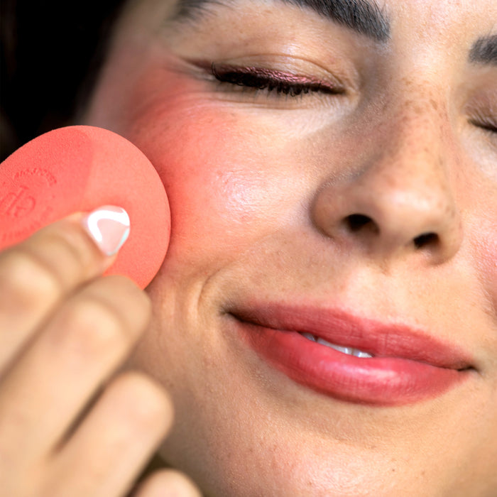 An expert uses the Ere Perez Bio All-Beauty Sponge to apply blush, achieving a seamless finish. Their skin glows with a rosy hue as they smile softly, eyes closed and lips in pink, embracing sustainable beauty practices.