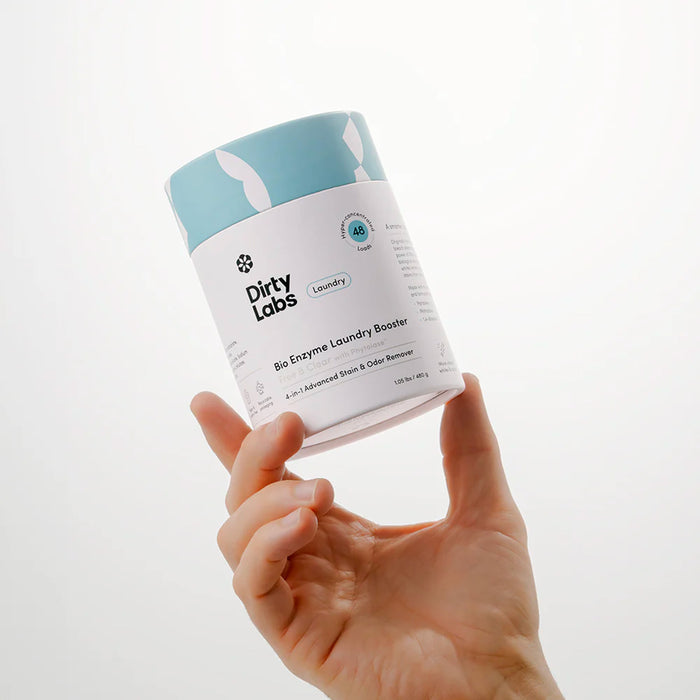 A hand holds a container of Dirty Labs Bio Enzyme Laundry Booster against a plain white background. The label, designed in blue and white, displays the product name and brand, emphasizing its advanced enzyme technology formulated to tackle biological stains.