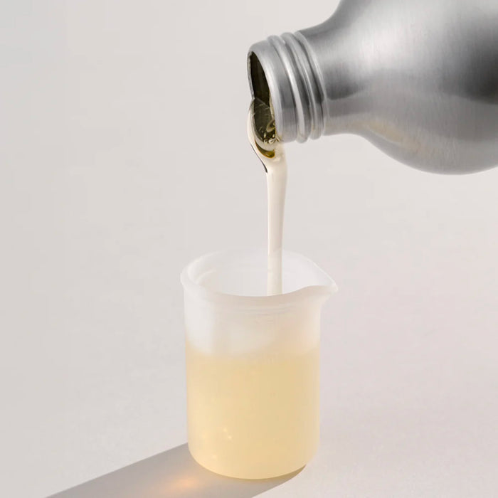 A metallic bottle of Dirty Labs Bio Laundry Detergent with Phytolase® - Free & Clear pours a clear, pale yellow liquid into a small, translucent plastic measuring cup on a light surface.
