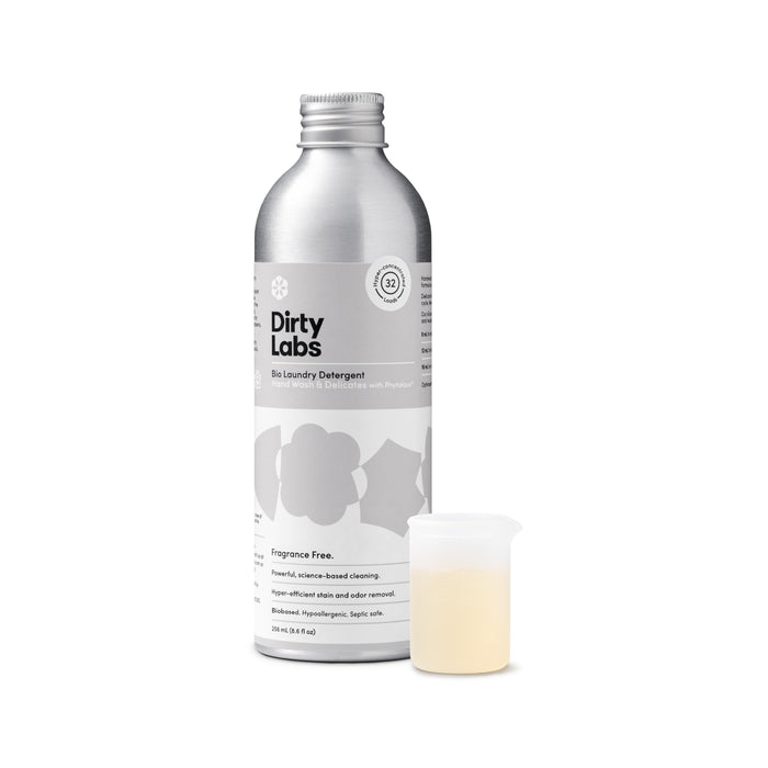 A silver bottle labeled Dirty Labs Bio Laundry Detergent with Phytolase® – Hand Wash & Delicates stands next to a small, clear measuring cup filled with light yellow liquid. This detergent is fragrance-free and suitable for sensitive skin.