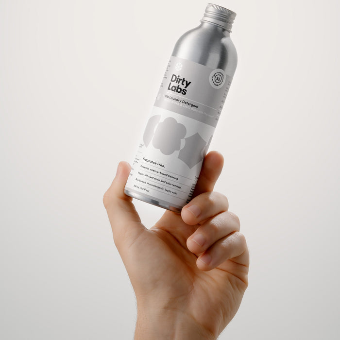 A hand holds a silver bottle labeled Dirty Labs Bio Laundry Detergent with Phytolase® – Hand Wash & Delicates, an enzyme-powered formula designed for effective yet gentle cleaning, against a plain white background. The minimalist bottle features sleek text and subtle graphics.