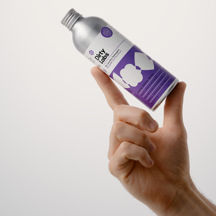 A hand holds up a silver bottle labeled Dirty Labs Bio Laundry Detergent with Phytolase® – Jasmine, Matcha, Vetiver, featuring a purple and white design, against a plain, light background.