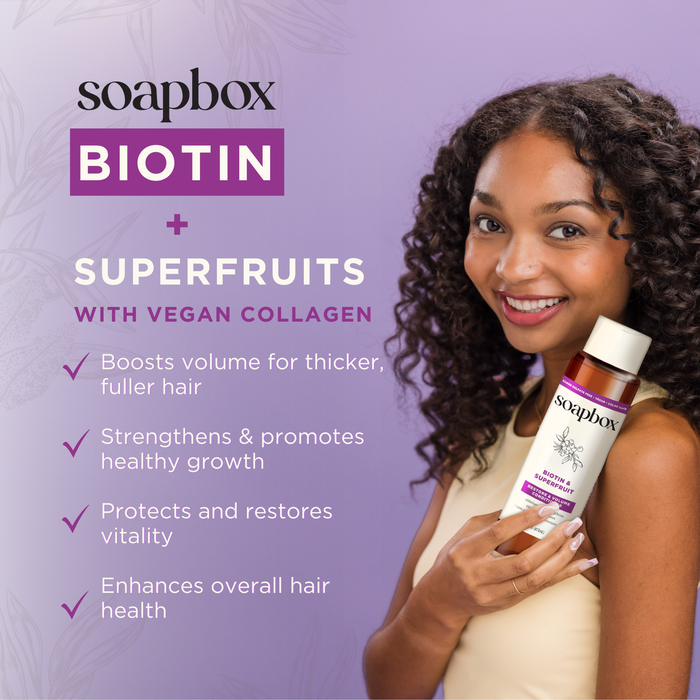 A person with curly hair beams while holding a bottle of Soapbox Biotin shampoo and the Biotin and Superfruit Restore & Volumize Conditioner, as the purple background highlights benefits like boosting volume, strengthening growth, protecting vitality, and enhancing hair health with vegan collagen.