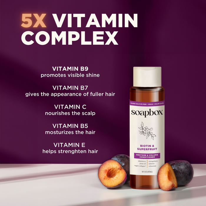 A bottle of Soapbox Biotin and Superfruit Restore & Volumize Conditioner sits on a surface. Surrounding text highlights its benefits: vitamin B9 for shine, B7 for fuller hair, C for scalp health, B5 for moisture, and E for strength. Two halved plums rest beside the bottle.