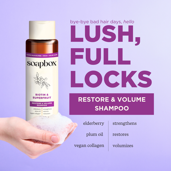 A hand holds a bottle of Soapbox Biotin and Superfruit Restore & Volumize Shampoo, foamy on the palm. The purple text reads Lush, Full Locks. This volumizing shampoo highlights its key ingredients. Background transitions from lavender to light purple.