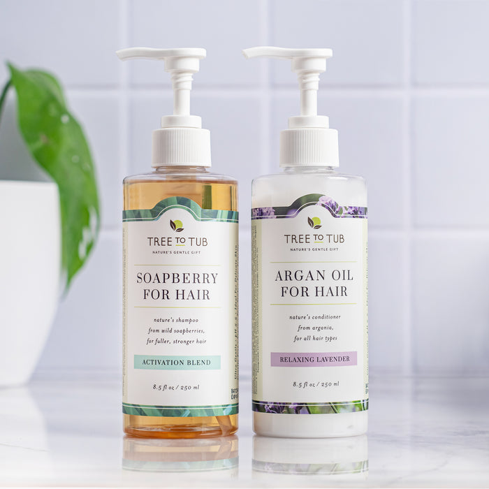 Two bottles of Tree To Tub hair products sit on a white surface. On the left is Biotin & Caffeine Volumizing Shampoo for Sensitive Scalp, and on the right is Argan Oil Conditioner. A green leaf in the background adds a natural touch to this hair loss prevention duo.
