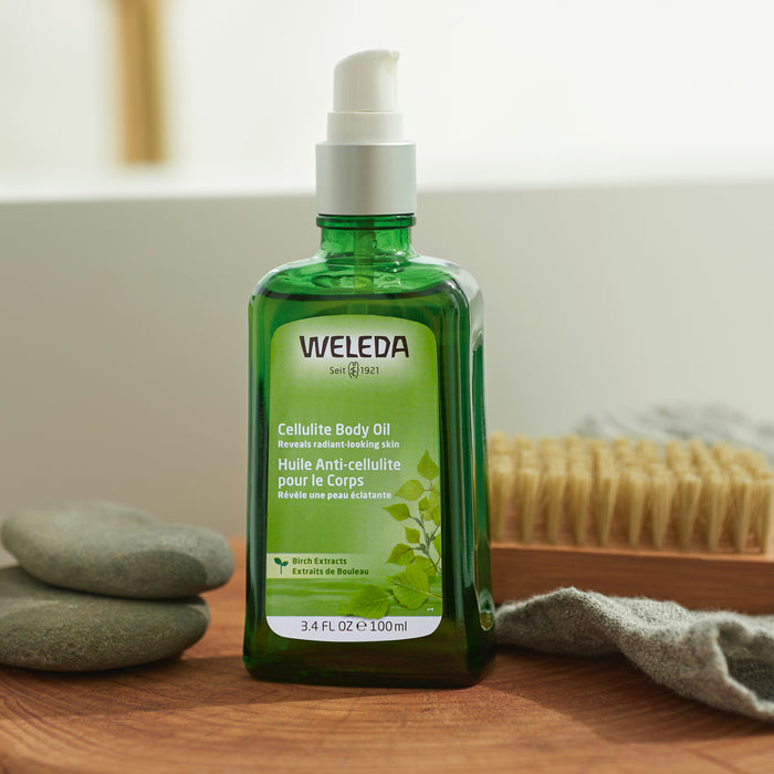 A green glass bottle of Weledas Birch Cellulite Body Oil with a white pump dispenser sits on a wooden surface, surrounded by smooth gray stones, a natural bristle brush, and a folded gray towel, creating an ideal spa-like setting for cellulite reduction.