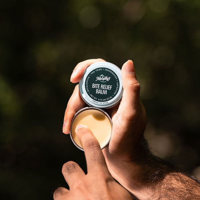 A person holds an open tin of Murphys Naturals Bite Relief Balm, dipping a finger into the light-colored balm inside. The green-labeled tin is ideal for your home pest control kit, ensuring relief is always within reach. The background remains out of focus.
