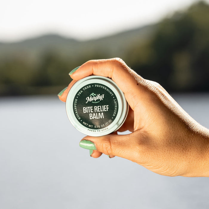A hand with green nails holds a Murphys Naturals Bite Relief Balm Tin against a blurred outdoor background of water and trees. The balm provides itch and discomfort relief from bites, offering a natural alternative to traditional pest control methods.