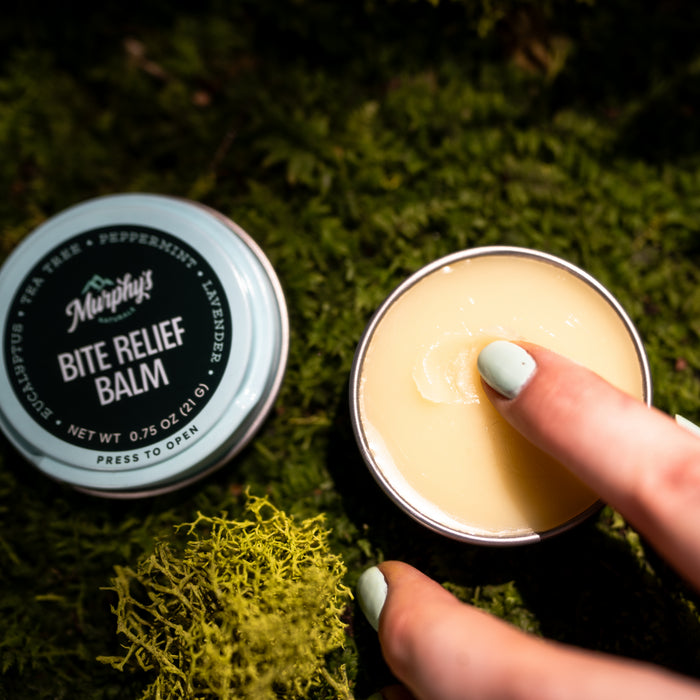 With mint green nail polish, a person dips a finger into an open tin of Murphys Naturals Bite Relief Balm. The tin sits on green moss against a brightly illuminated background, enhancing the serene setting.