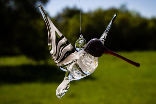 Hanging glass hummingbird with black and white swirl wings against a green outdoor background.